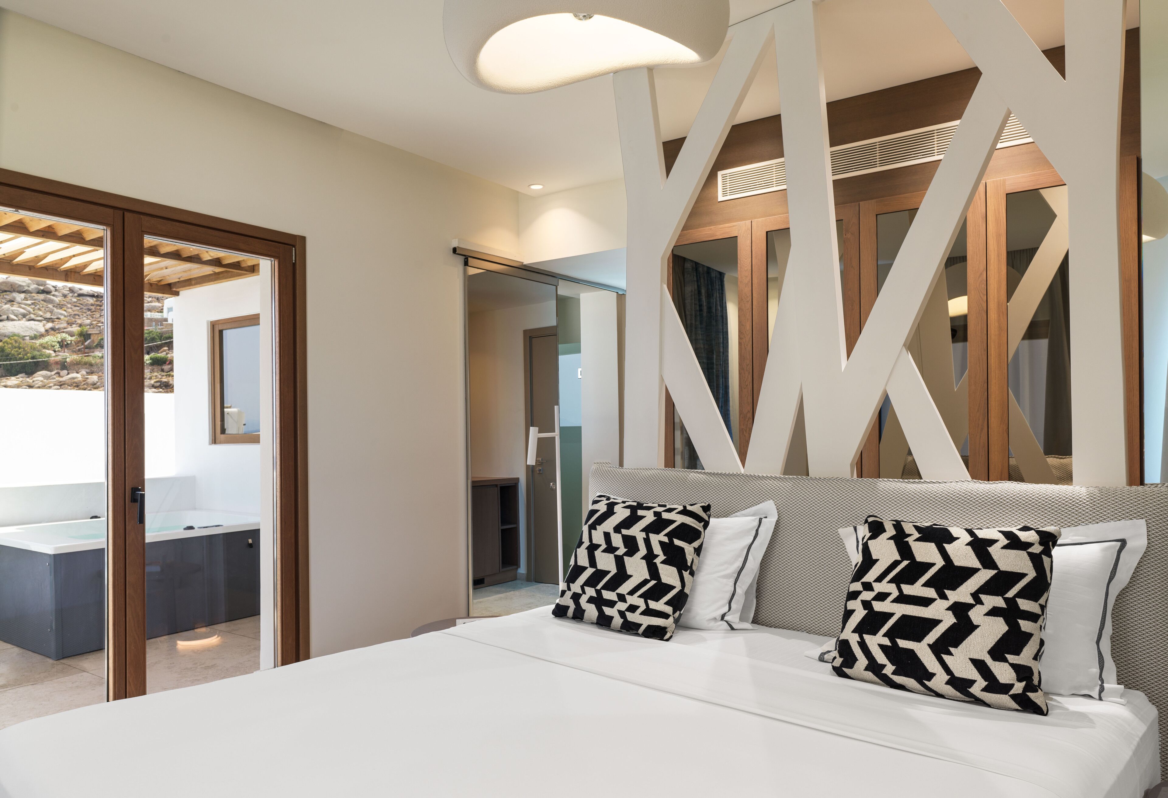 Une chambre moderne avec un grand lit, deux coussins à motifs, une tête de lit ornée de motifs géométriques et une vue sur la salle de bain et l’espace extérieur, visible à travers une porte en verre.
