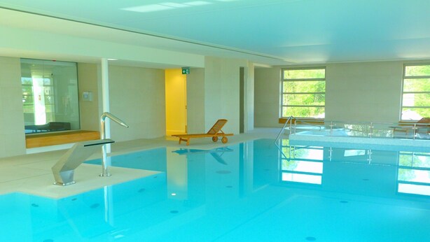 Une piscine intérieure à l’eau d’un bleu limpide, une pièce baignée de lumière grâce à de grandes fenêtres, et une chaise en bois près de la piscine.