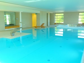 An indoor swimming pool with clear blue water, a sunlit room with large windows, and a wooden chair near the pool. - Zamek Biskupi Janów Podlaski (Janow Podlaski)