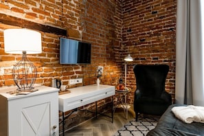 A room with a brick wall, a flat-screen TV mounted above a white desk, a black chair, and a bed with a white blanket. - Zamek Biskupi Janów Podlaski (Janow Podlaski)