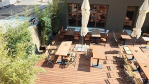 Un patio al aire libre con mesas y sillas de madera, algunas con sombrillas, y plantas en macetas.