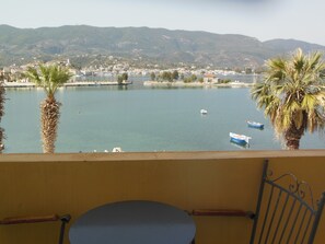 A balcony with a view of a calm lake, boats, and a distant town. - Theano Hotel (Poros)