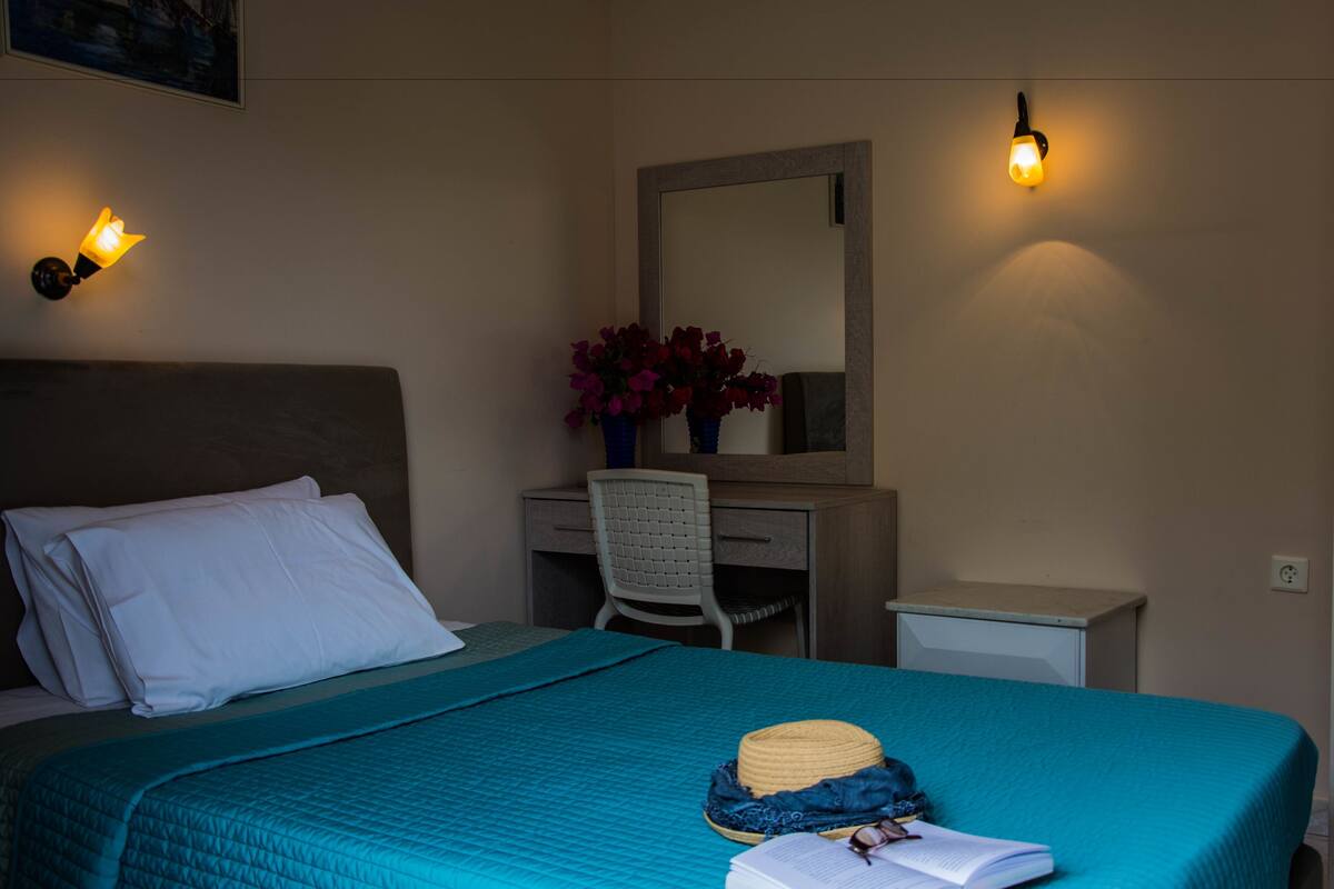 A neatly made bed with a teal bedspread, a white pillow, and a hat on the bed. There is a desk with a chair, a mirror, and a vase with flowers. Two wall sconces provide lighting.