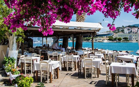 An outdoor dining area with tables and chairs, overlooking a body of water and a hillside town.