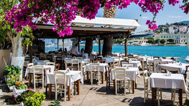 Area pranzo all'aperto con tavoli e sedie, affacciata su un corpo d'acqua e una cittadina collinare.