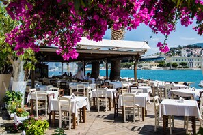 An outdoor dining area with tables and chairs, overlooking a body of water and a hillside town. - Theano Hotel (Poros)