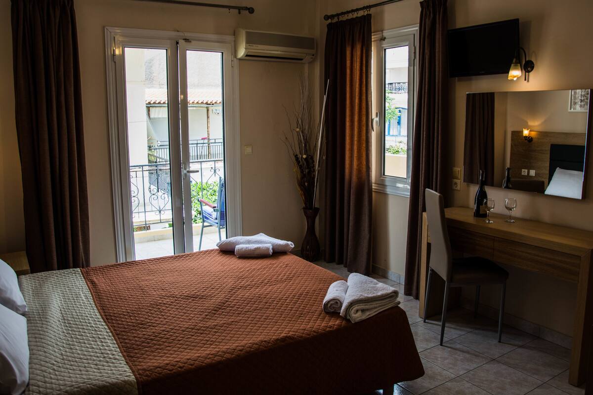 A hotel room with a bed, a desk, a chair, a TV, and a balcony with a railing.
