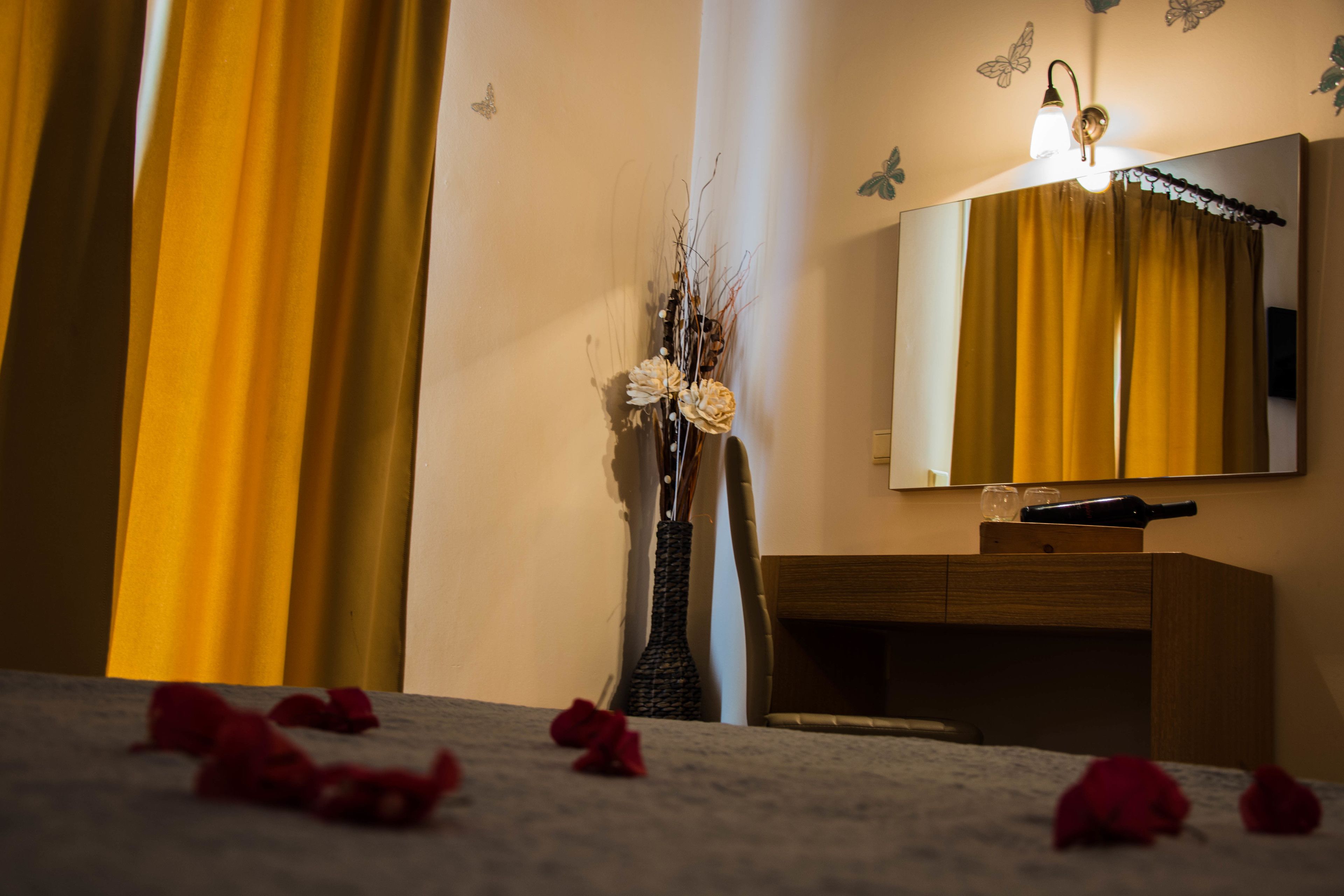 A hotel room with a bed, a wooden dresser, a vase with flowers, and a mirror with curtains.