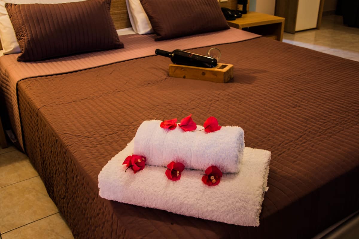 A neatly made bed with a brown bedspread, two white towels with red flowers, and a bottle on a wooden tray.