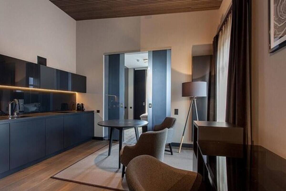 Una cocina moderna con muebles oscuros, una mesa redonda para comer y un sofá. Hay una lámpara y se ve el pasillo.