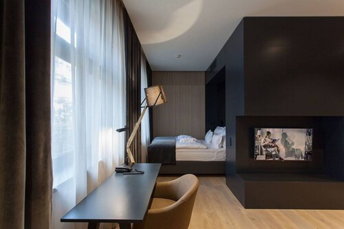 Una habitación de hotel moderna con cama, escritorio, silla, televisor y un amplio ventanal con cortinas.