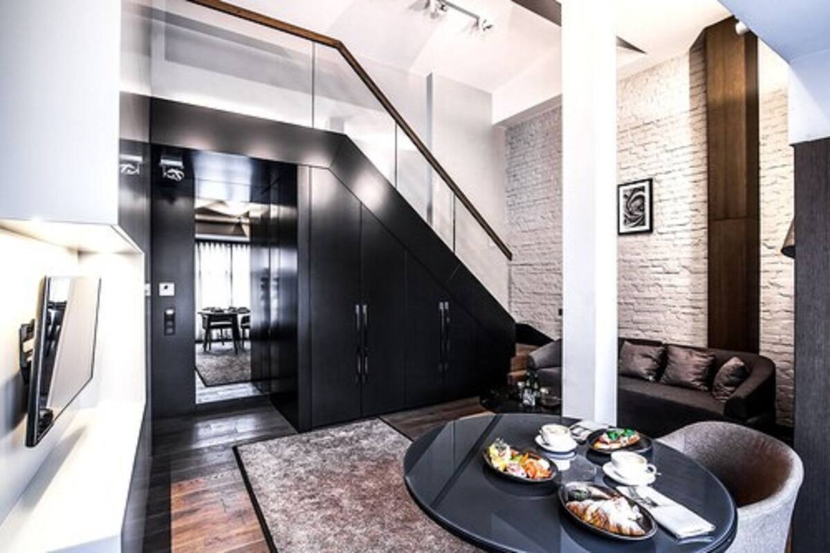 Un salón moderno con una escalera, piso de madera oscura, una mesa redonda con comida, un sofá y una pared de ladrillo.