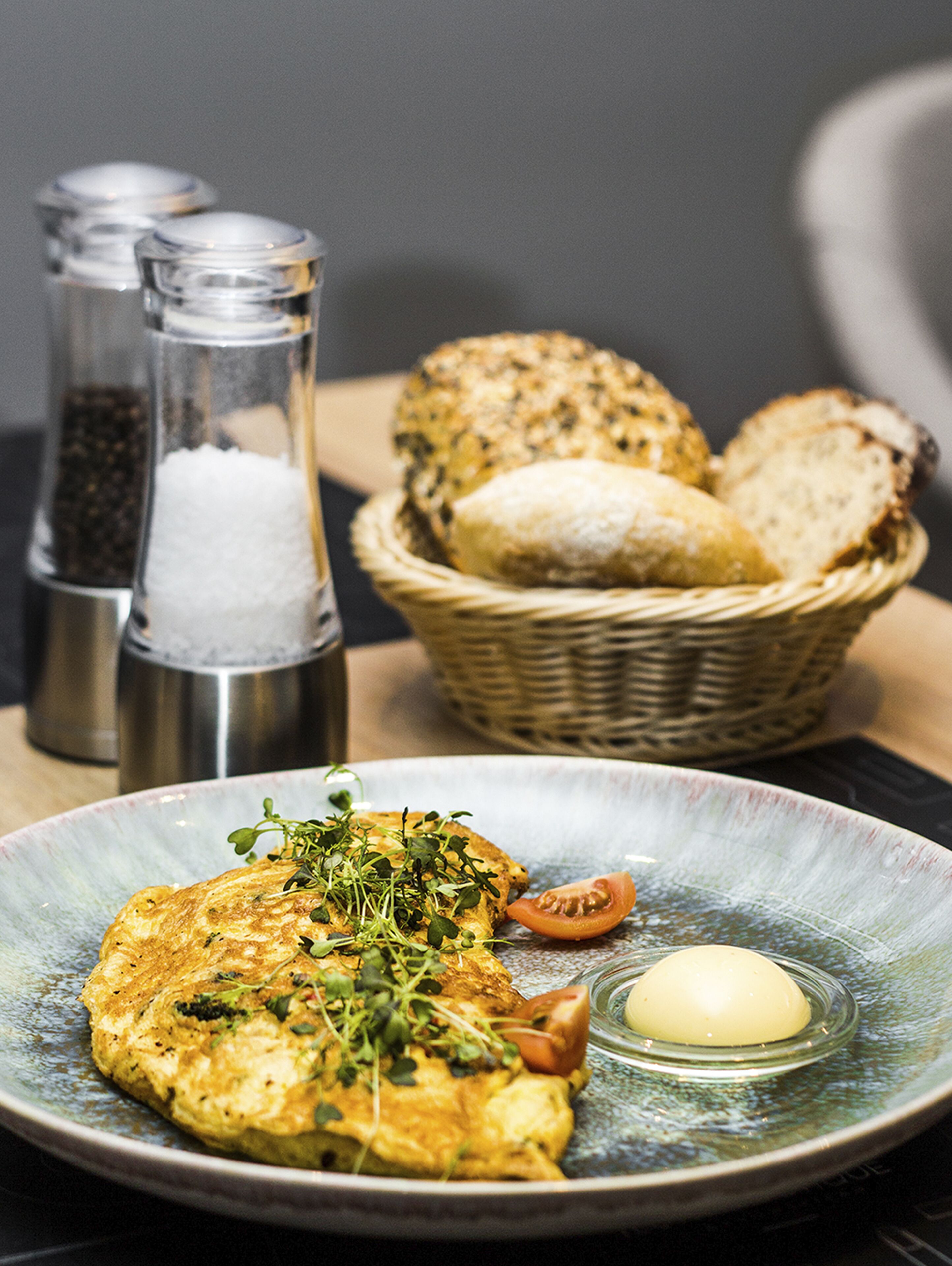 Ein Teller mit einem Omelett, einer Tomatenscheibe und einem Klecks Butter. Im Hintergrund stehen Salz- und Pfefferstreuer sowie ein Brotkorb.