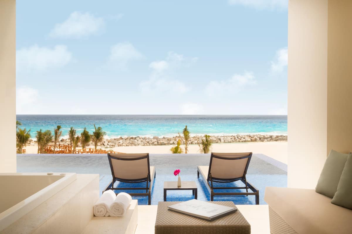 A balcony with a view of the ocean, two chairs, a small table with a vase, and a bathtub.