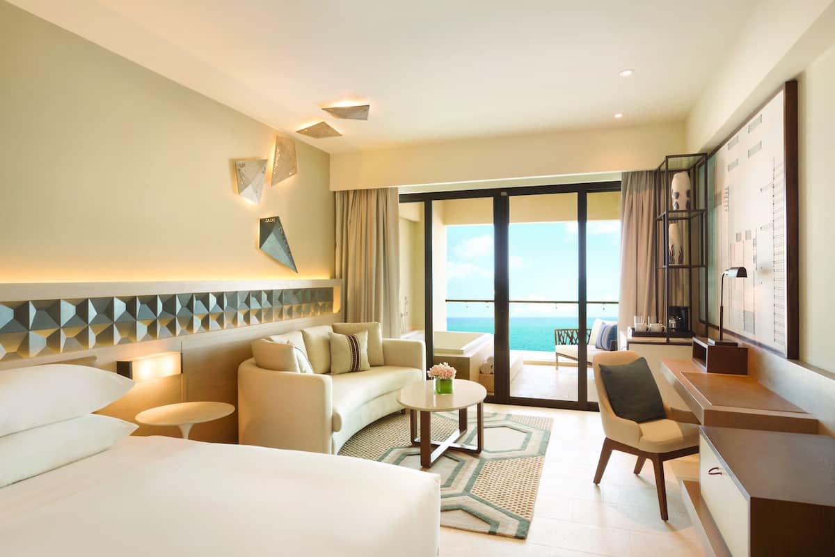 A hotel room with a bed, a sofa, a chair, a small table, and a desk. There is a large window with a view of the ocean.