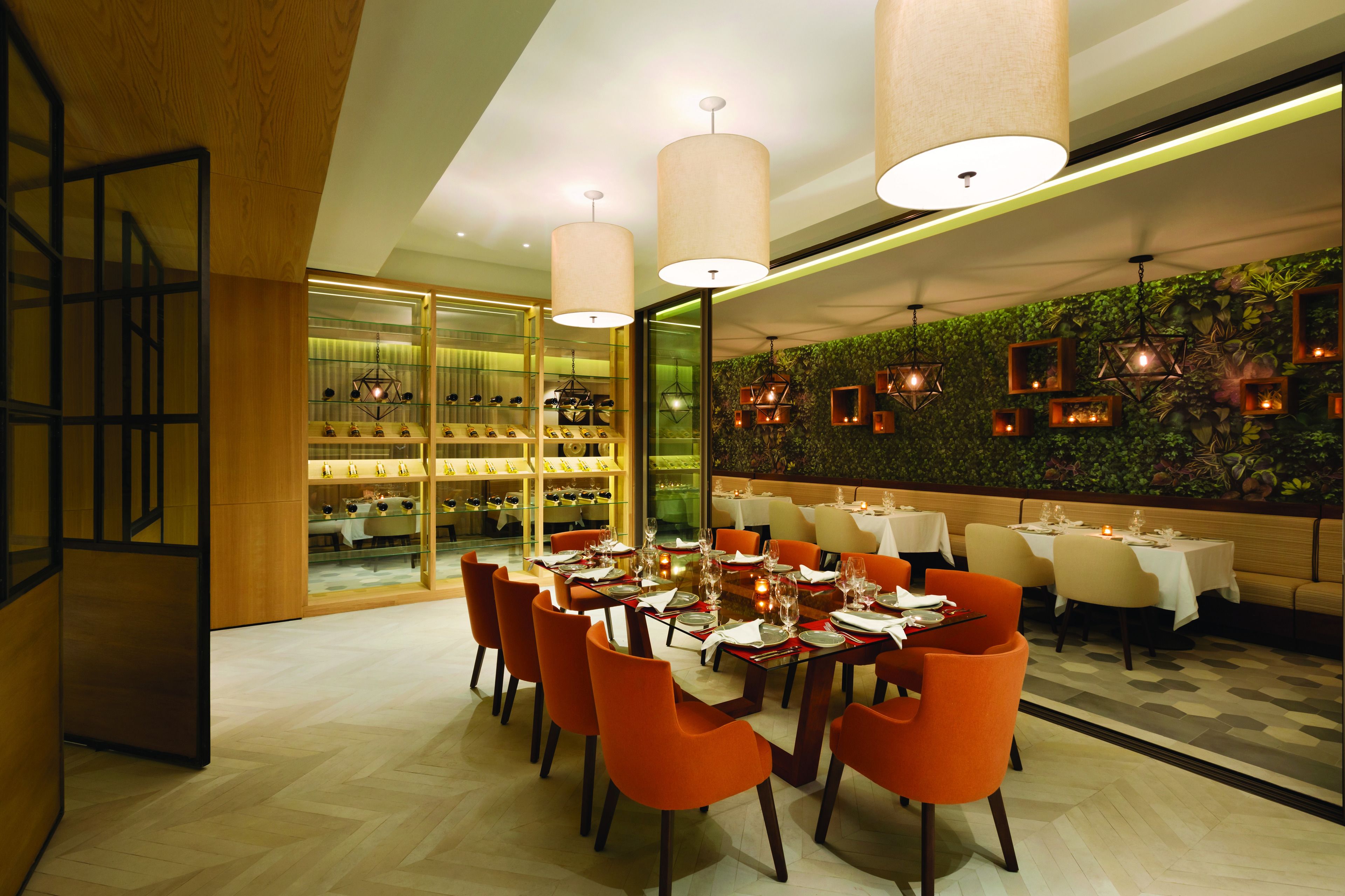 A modern restaurant with a long dining table, orange chairs, and a green wall with plants.