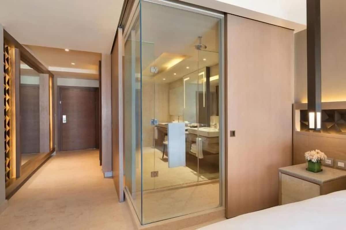 A modern hotel room with a glass partition, a bed, a bedside table with a vase, and a headboard with geometric lighting.