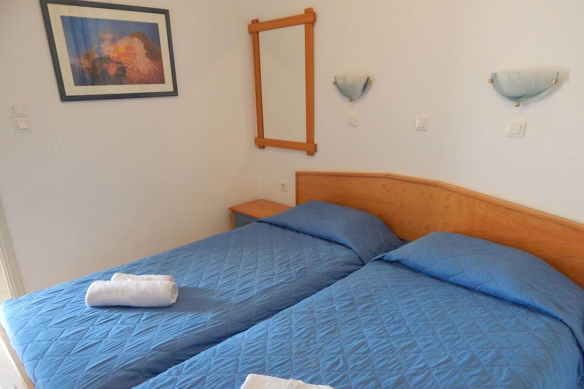 A bedroom with a wooden headboard, two blue bedsheets, a white towel, a nightstand, a framed picture, and two wall-mounted light fixtures.