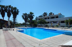 En swimmingpool med solstolar och parasoller, omgiven av palmer och en byggnad med flera våningar.