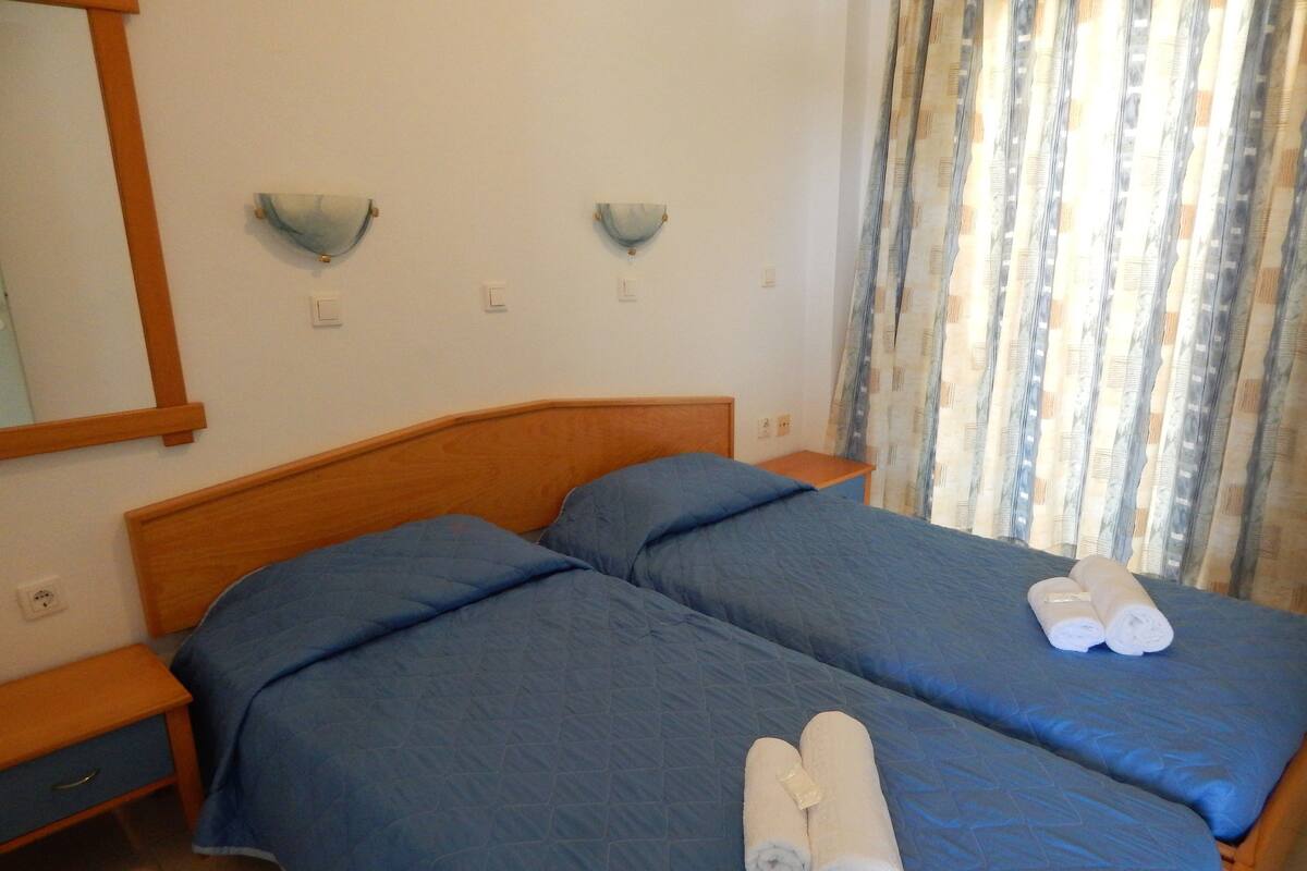 A bedroom with two beds, each with blue bedding and a wooden headboard. There are two wall-mounted nightstands and a mirror on the left wall.