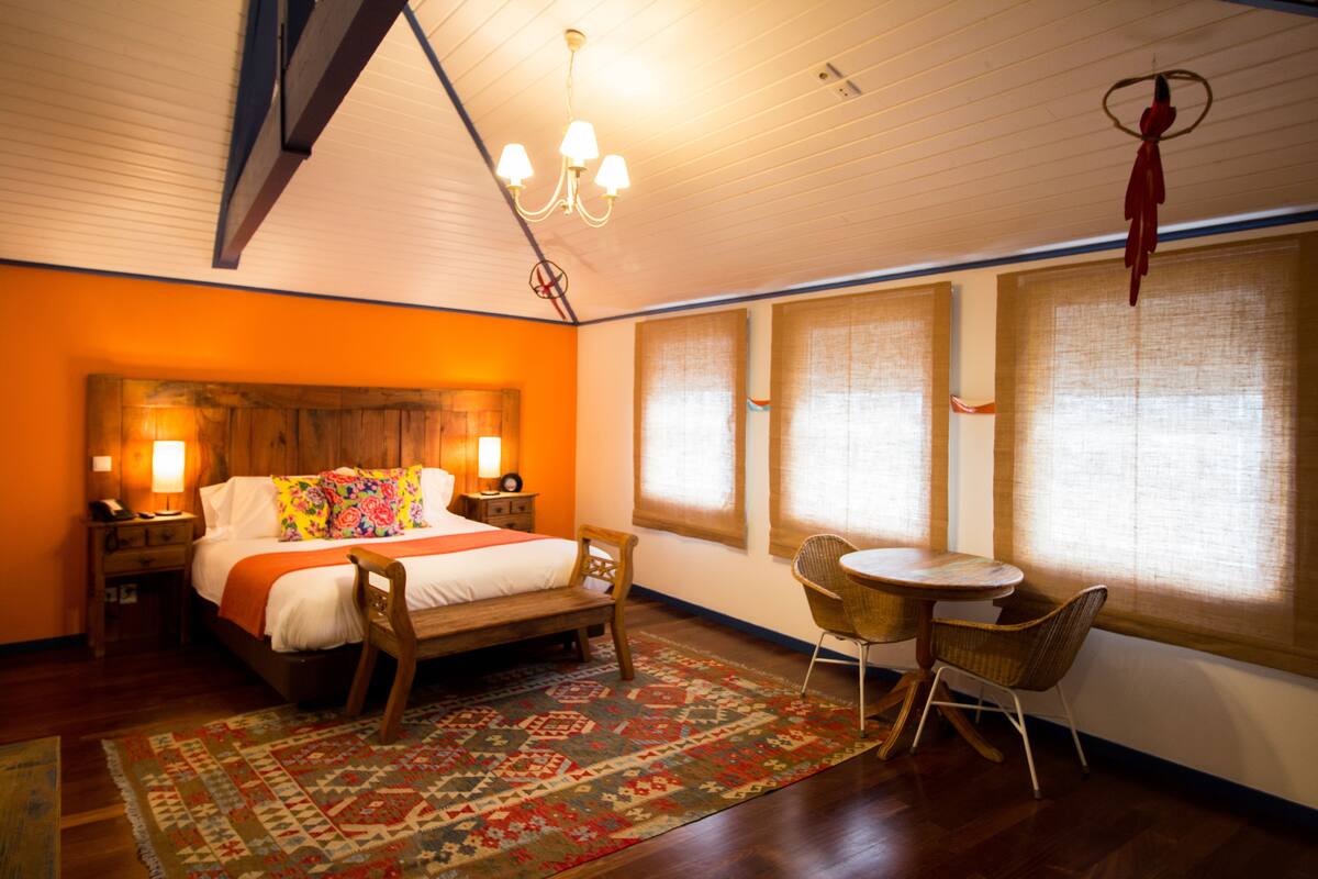 A hotel room with a wooden bed, a round table, and a chair. There are two windows with blinds, a chandelier, and a red hanging decoration.
