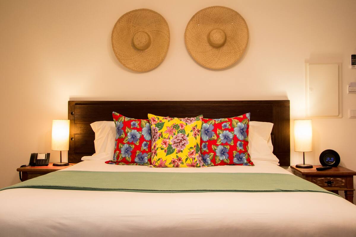 A bed with floral pillows, two wall hats, bedside lamps, a phone, and a clock on a wooden nightstand.