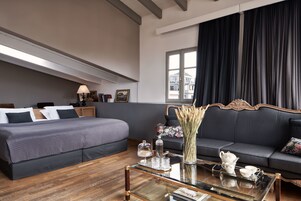 Moderni hotellihuone, jossa on suuri sänky, sohva, lasinen sohvapöytä ja ikkunasta avautuva näkymä kaupunkiin.