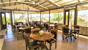 A covered outdoor dining area with multiple round tables and chairs, large windows, and a view of greenery and buildings.