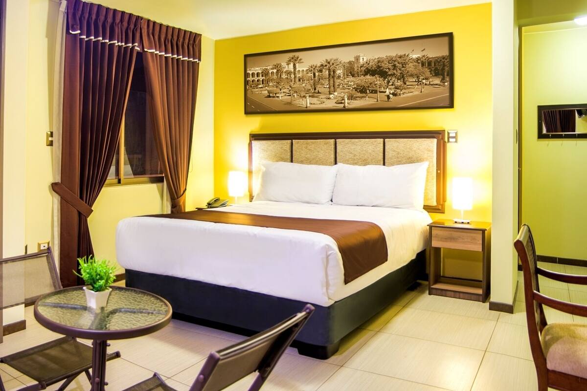 Une chambre d’hôtel avec un lit, des tables de chevet, un bureau, une chaise, une petite table avec une plante et une grande fenêtre avec des rideaux.