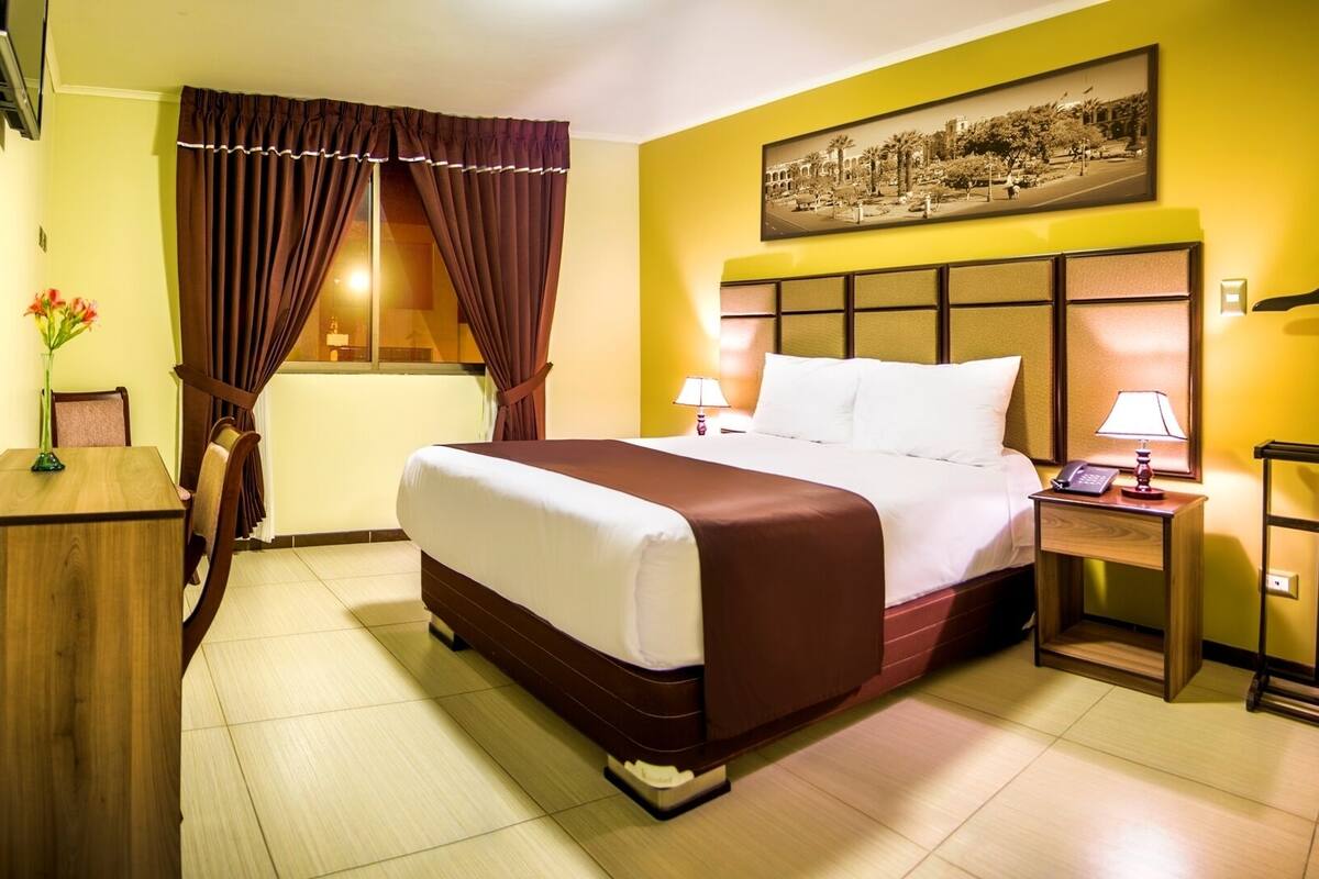 Une chambre d’hôtel avec un lit, des tables de chevet, une lampe, un tableau au mur et une fenêtre avec des rideaux.