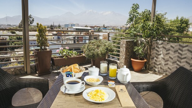 A breakfast setup on a balcony with a view of mountains, including a plate of scrambled eggs, coffee, juice, and a basket of bread.