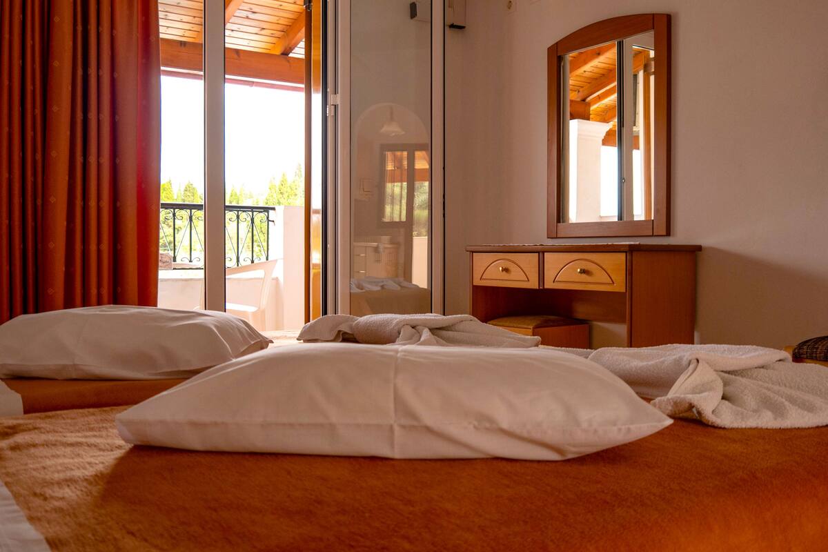 Una habitación de hotel con cama, cómoda de madera, espejo y balcón con cortinas rojas.