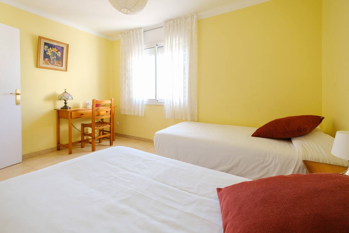 A room with two beds, a desk with a chair, a window with white curtains, and a painting on the wall.