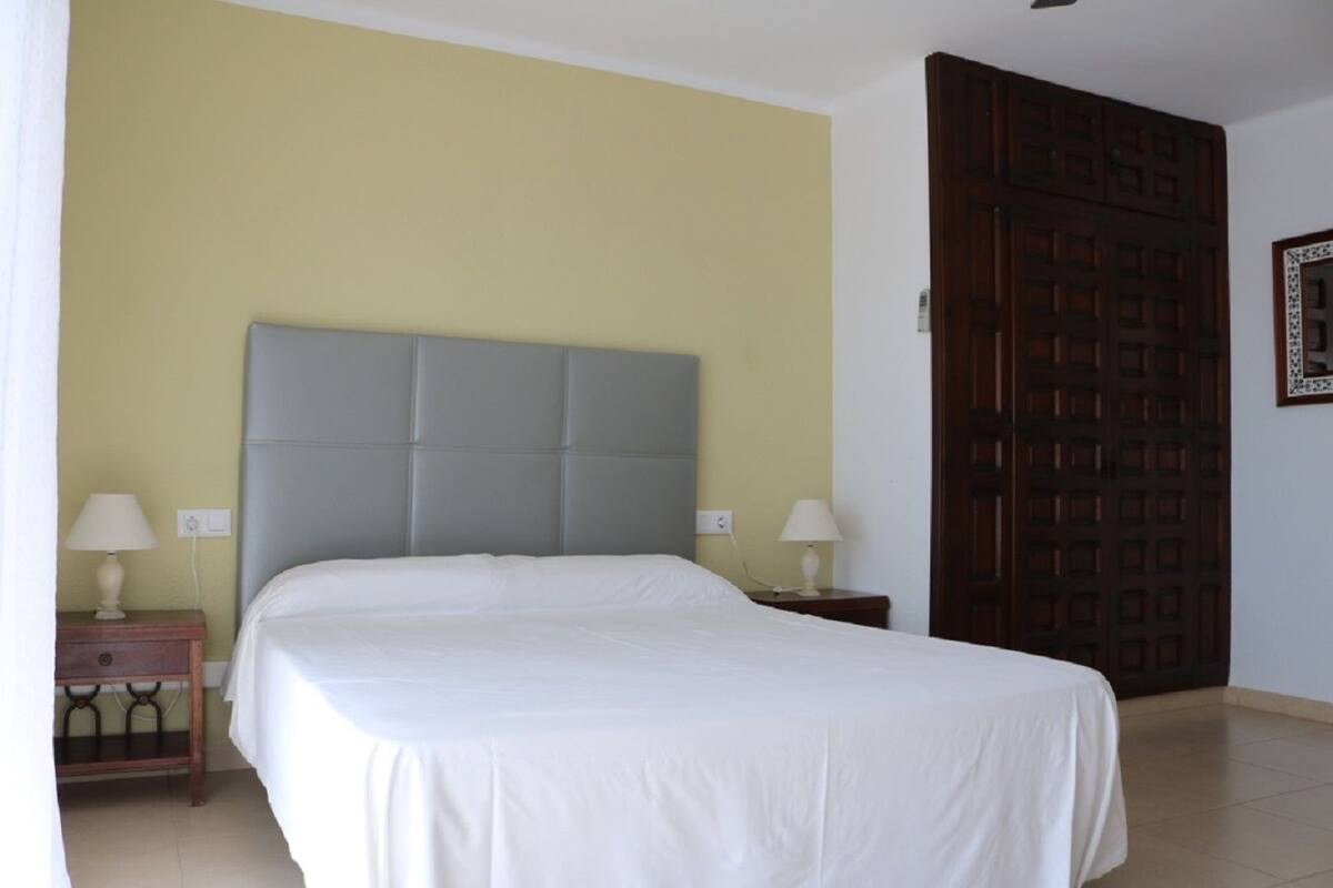 A bedroom with a bed, two bedside tables, a nightstand, a wooden door, and a wall-mounted light switch.