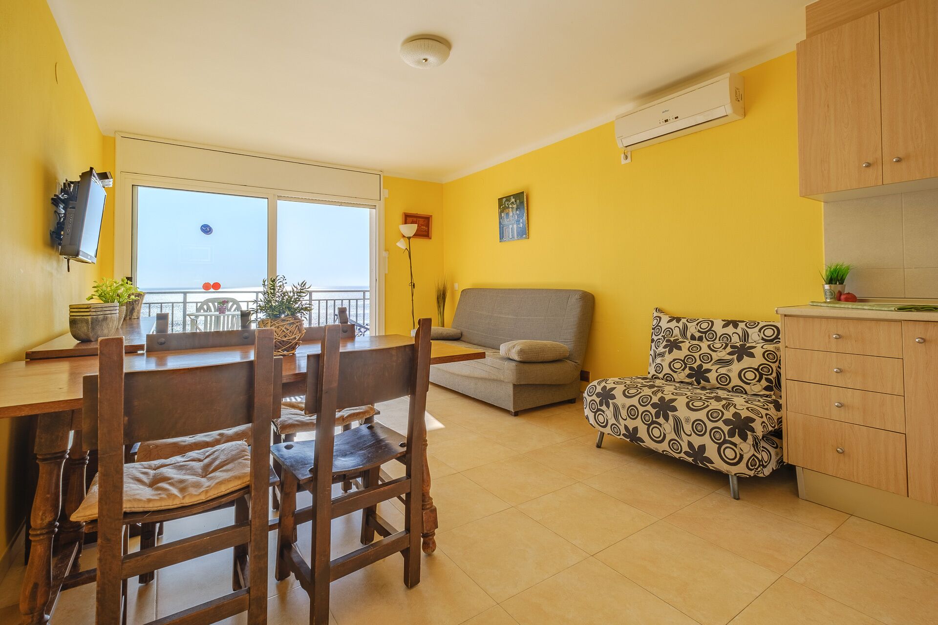 A bright yellow room with a dining table, chairs, a sofa, a patterned armchair, and a kitchen area with wooden cabinets.