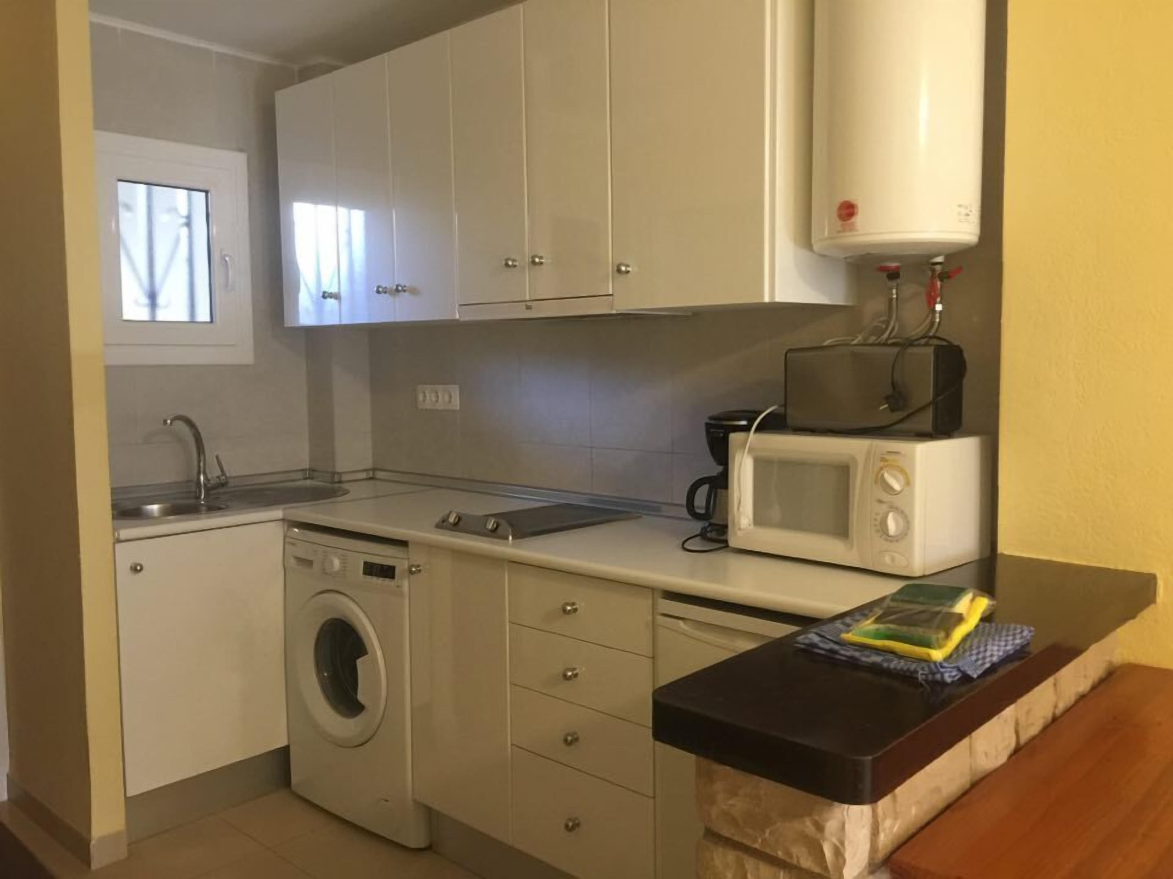 Una cocina compacta con armarios blancos, lavadora, microondas y cafetera.