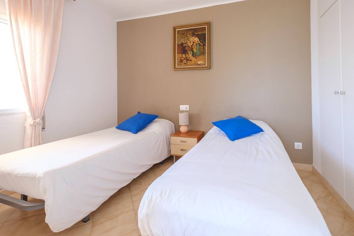A hotel room with two single beds, a nightstand, a lamp, and a painting on the wall.