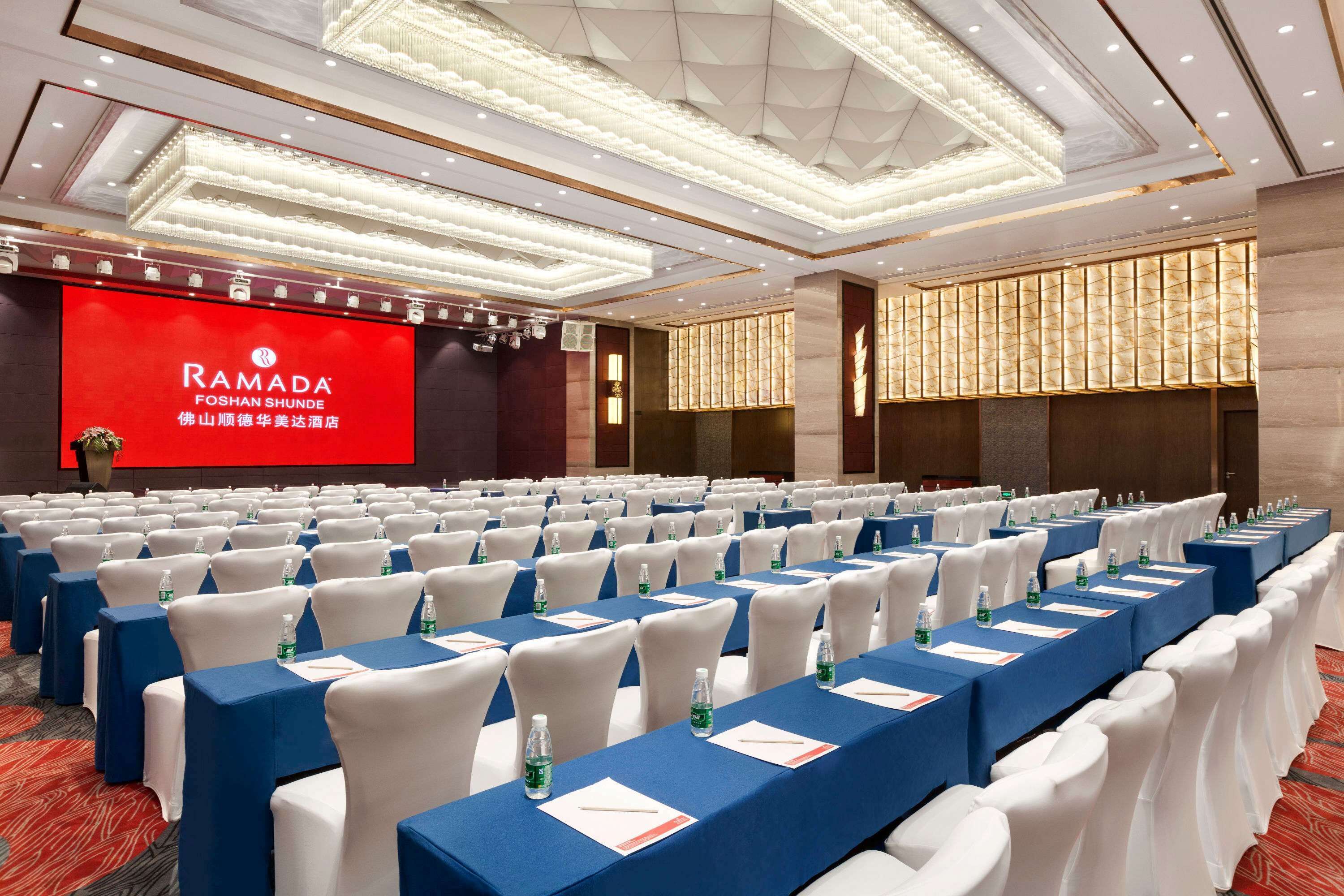 A conference room with rows of tables and chairs, a large screen displaying the Ramada logo, and a chandelier.