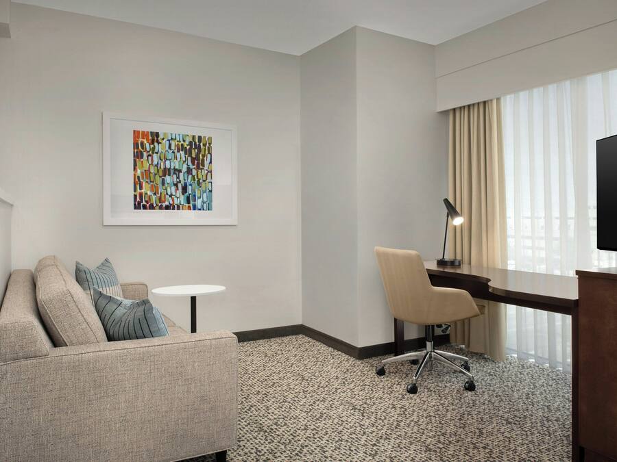 A hotel room with a sofa, a desk, a chair, a small table, a framed artwork, and a television.
