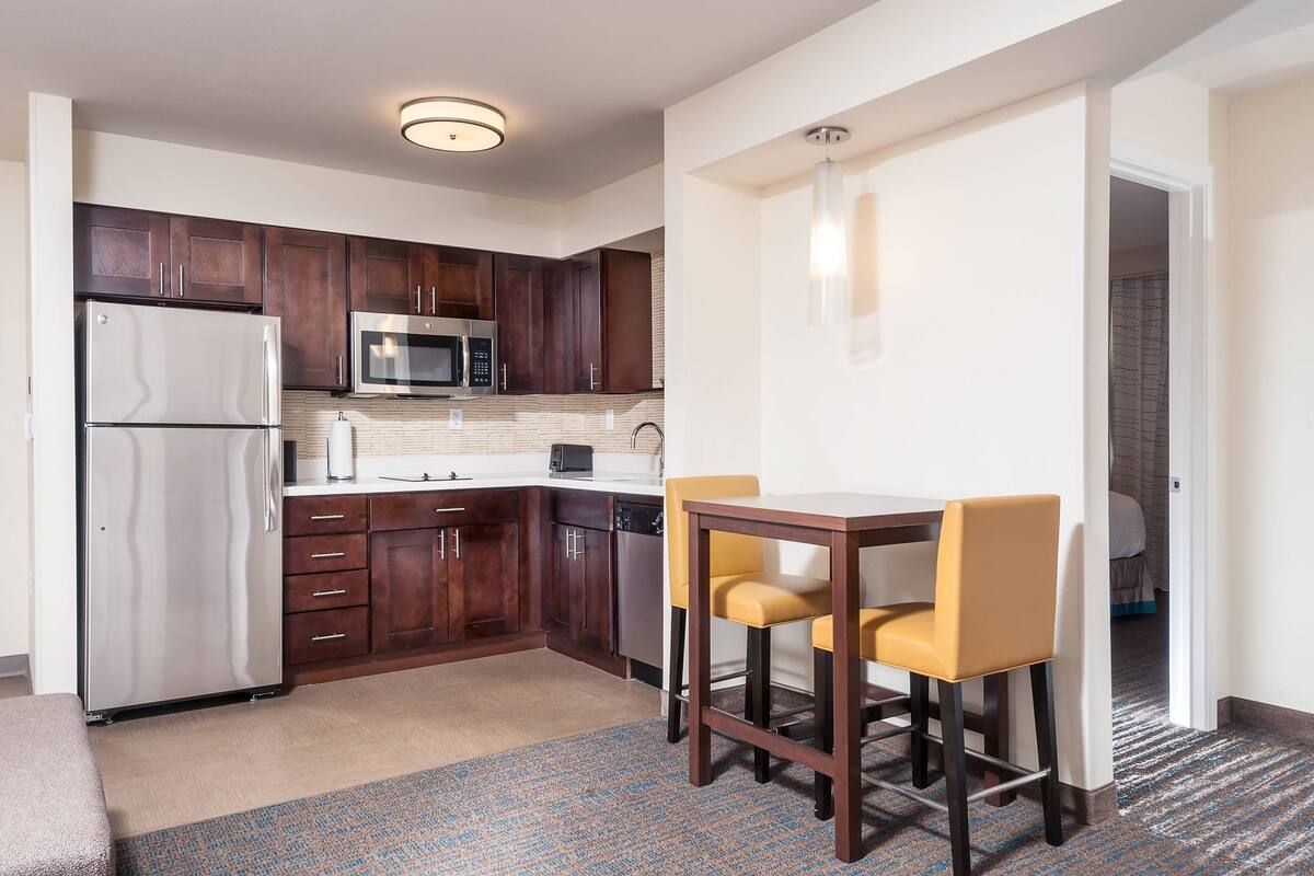 A compact kitchen with dark wood cabinets, a stainless steel refrigerator, and a microwave. There is a small dining area with two yellow chairs and a wooden table.