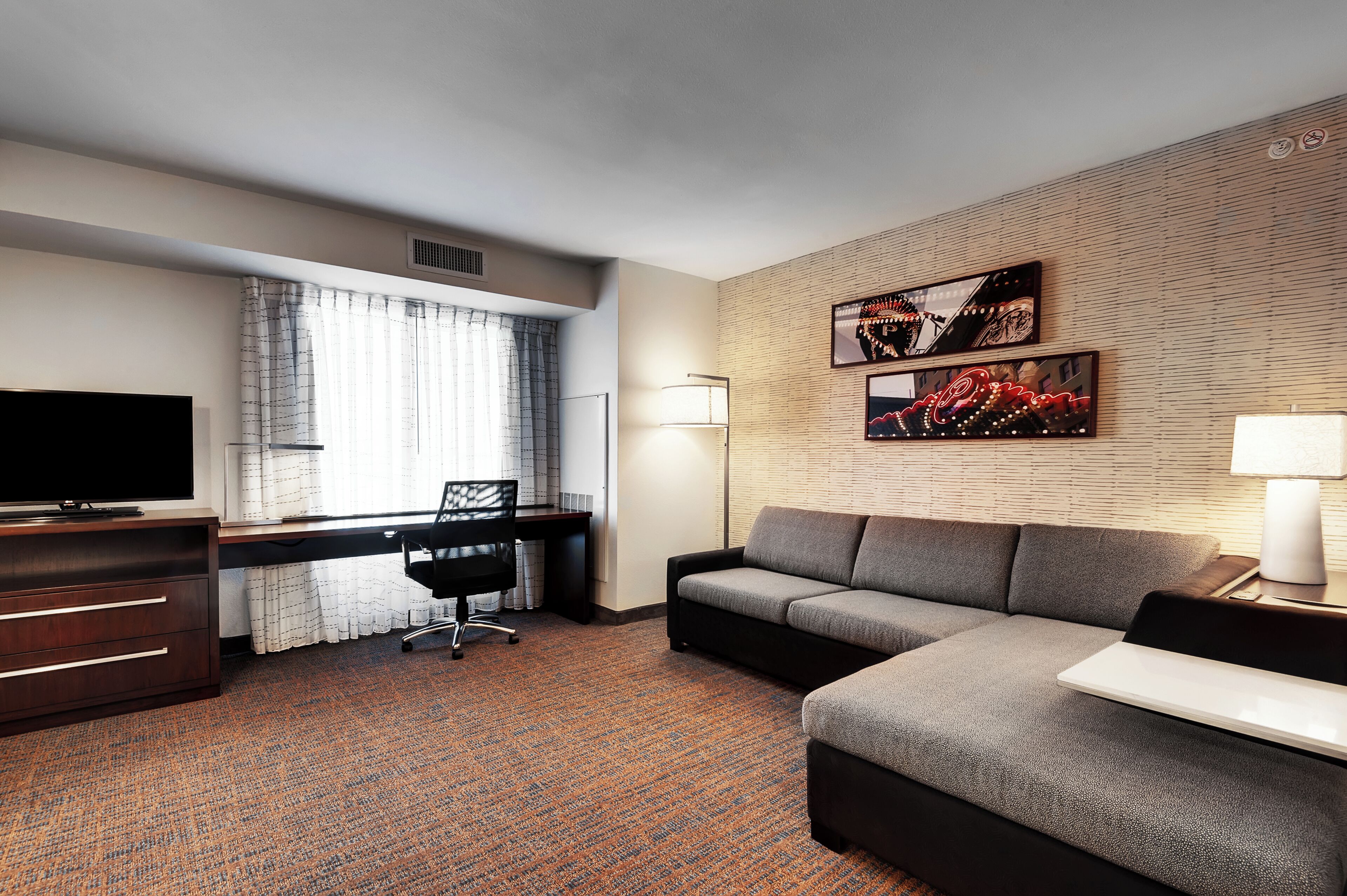 A hotel room with a sofa, a desk with a chair, a TV, and artwork on the wall.