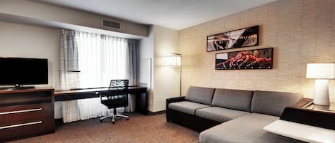 A hotel room with a sofa, a desk with a chair, a TV, and artwork on the wall.