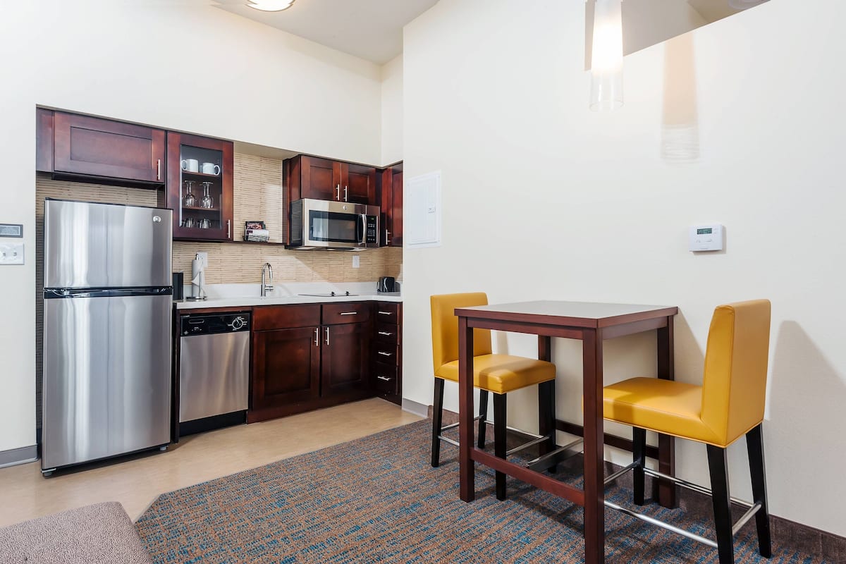A compact kitchen with a refrigerator, microwave, and dishwasher, a small dining table with two chairs, and a patterned rug.