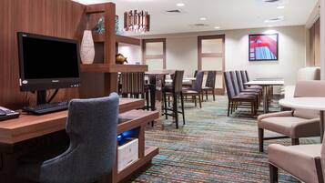 A hotel room with a large flat-screen TV, a desk with a keyboard, a printer, and a chair. There are multiple tables and chairs in the room, and a large window with a view of a building.