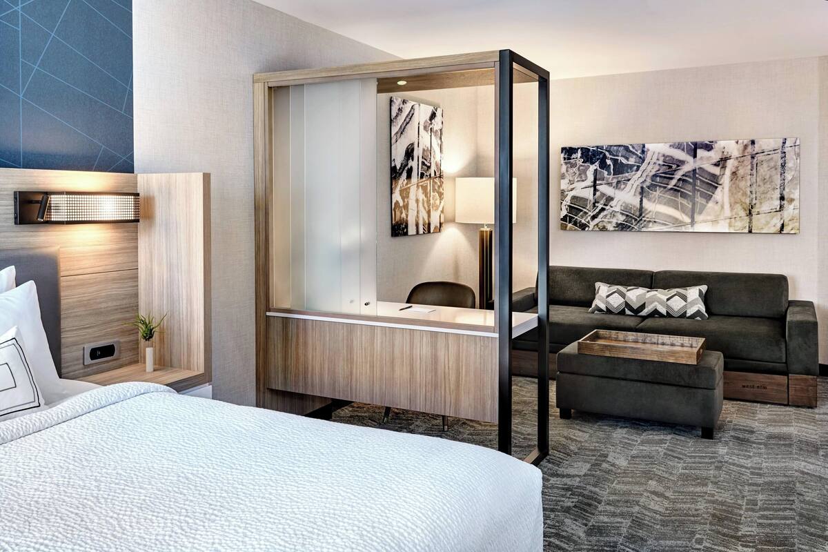 A hotel room with a bed, a desk with a chair, a sofa, a coffee table, and a large abstract painting on the wall.