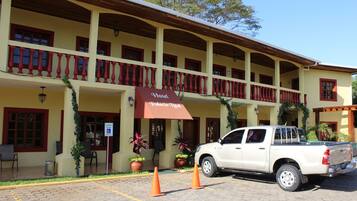 Un hotel con toldo rojo, mesas al aire libre y una camioneta pickup blanca estacionada adelante.