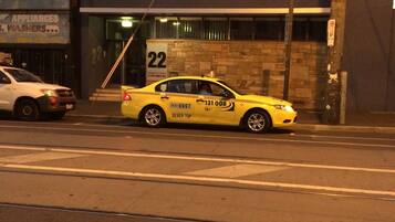 A yellow taxi with the number 22 and "TRAVELLERS ACCOMODATION" on the side, parked in front of a building with a sign that reads "ELECTRICAL APPLIANCES, WASHERS".