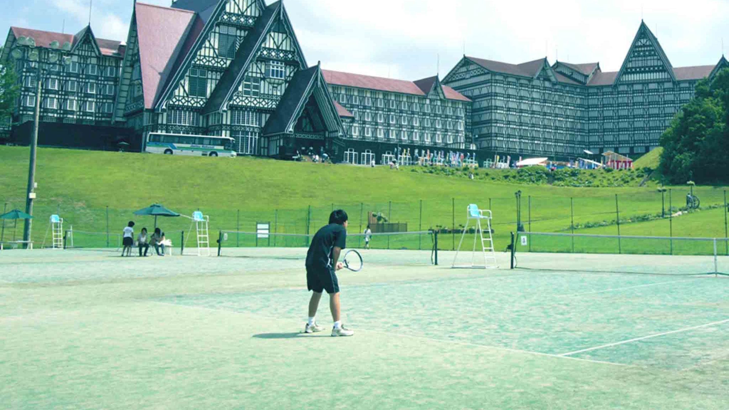 A tennis court with a player in action, surrounded by a large building with a steeply pitched roof and multiple windows.