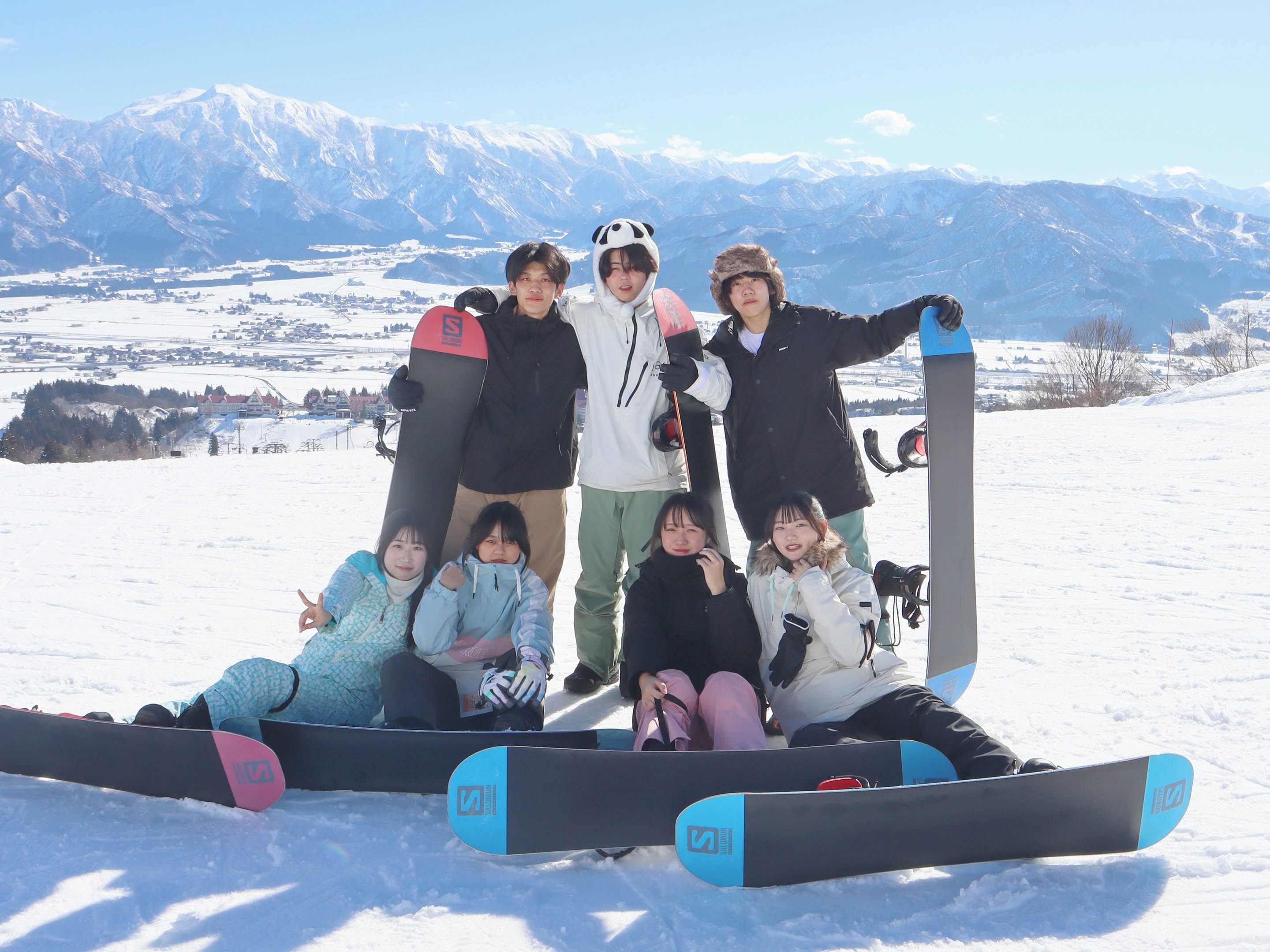 A group of people posing with snowboards on a snowy mountain with a clear view of distant mountains.