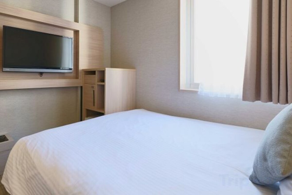 A hotel room with a bed, a television mounted on the wall, a bedside table, and a window with curtains.